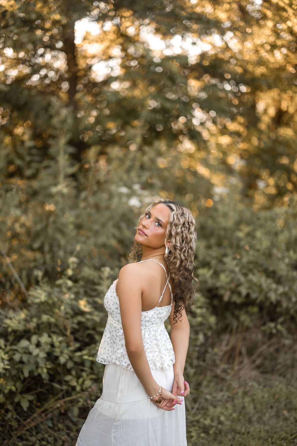 A dreamy outdoor portrait of a woman in a white lace dress poses amongst golden sunlit foliage at sunset.