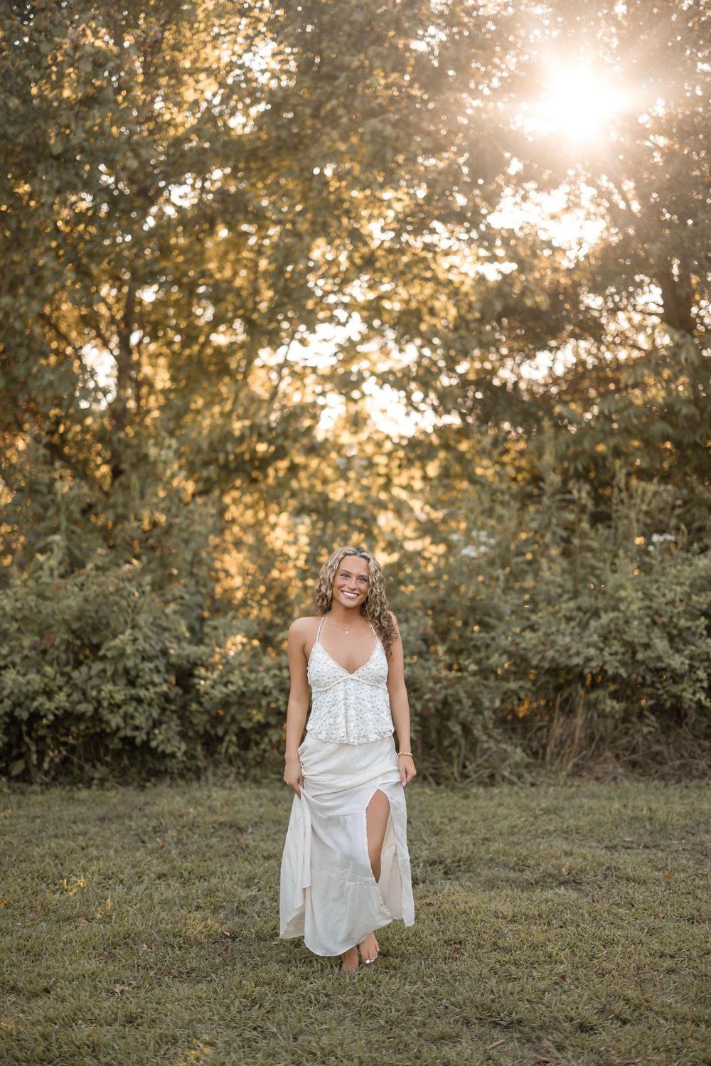 Sunlit figure in a flowing white dress stands gracefully in a grassy meadow at golden hour.