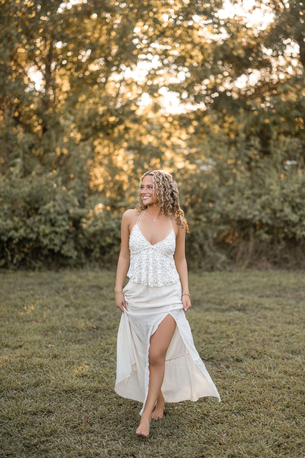 A person in a flowing white dress sits gracefully on grassy ground during golden hour sunset.