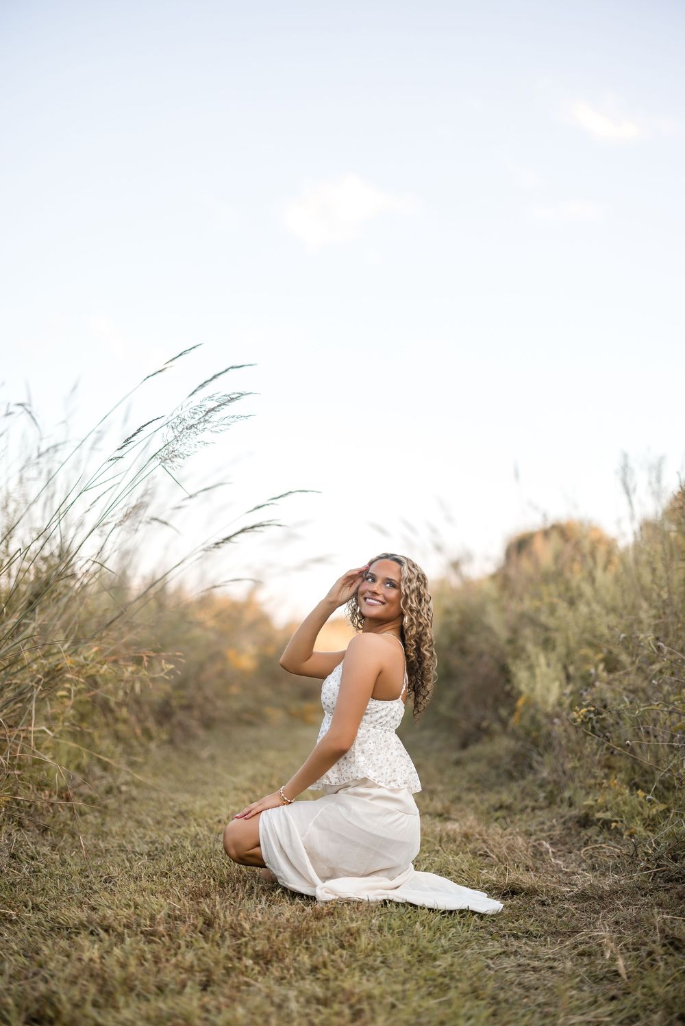 Woman in flowing white dress sits gracefully in tall grass during a golden hour photoshoot.
