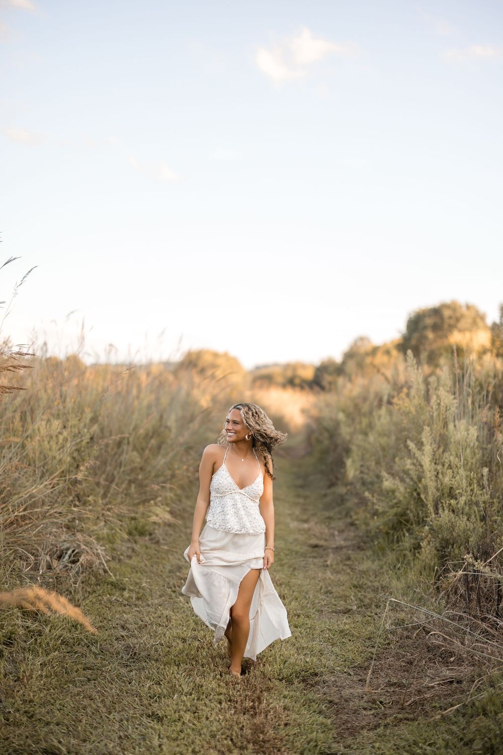 A flowing white dress catches the golden sunlight on a rustic dirt path through tall grasses at sunset.