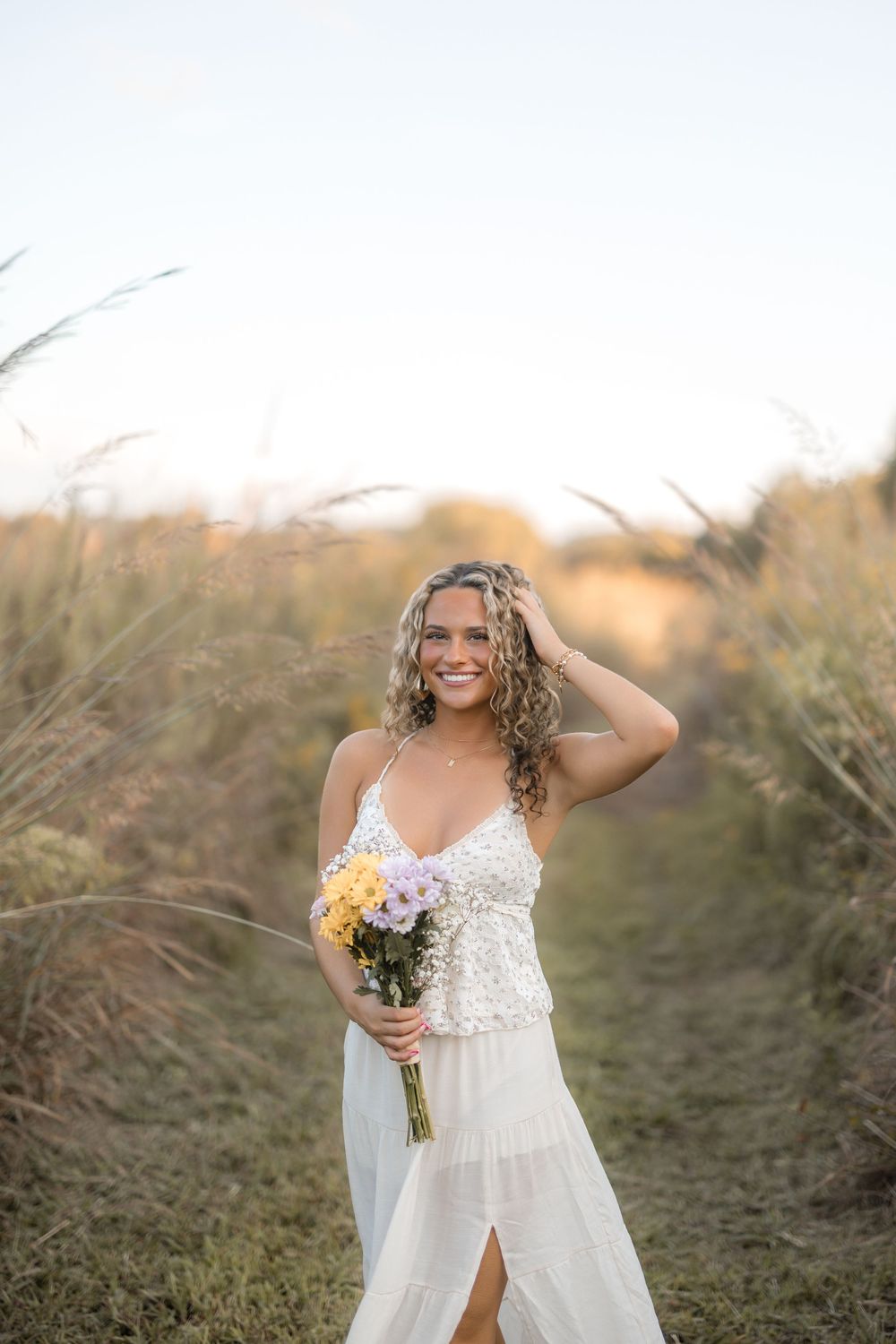 A bride in a white lace dress poses with a yellow bouquet against a natural outdoor backdrop at sunset.