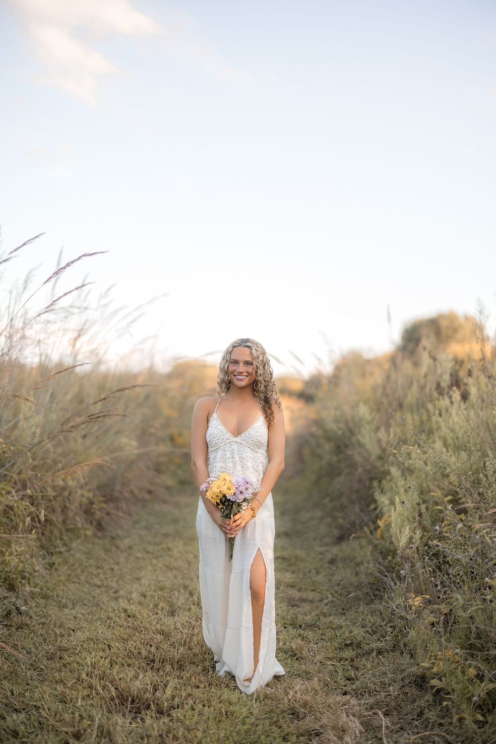 A white flowy dress and colorful wildflower bouquet stand out against a natural field setting at dusk.