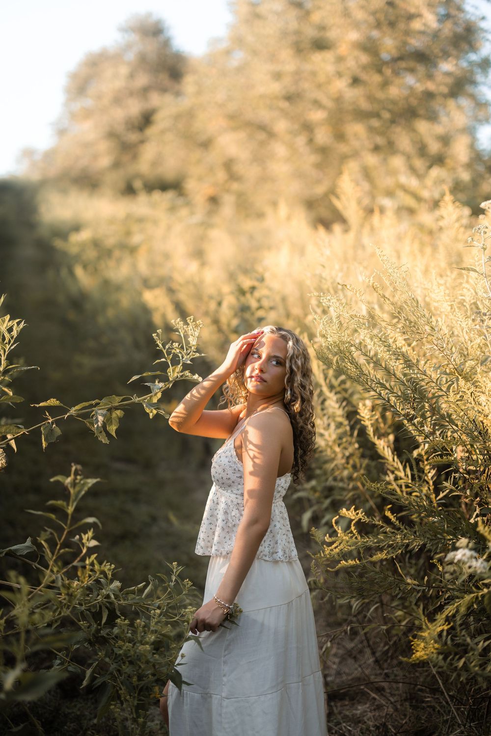 A white floral dress stands out against tall grass and golden sunlight in a dreamy summer field portrait.