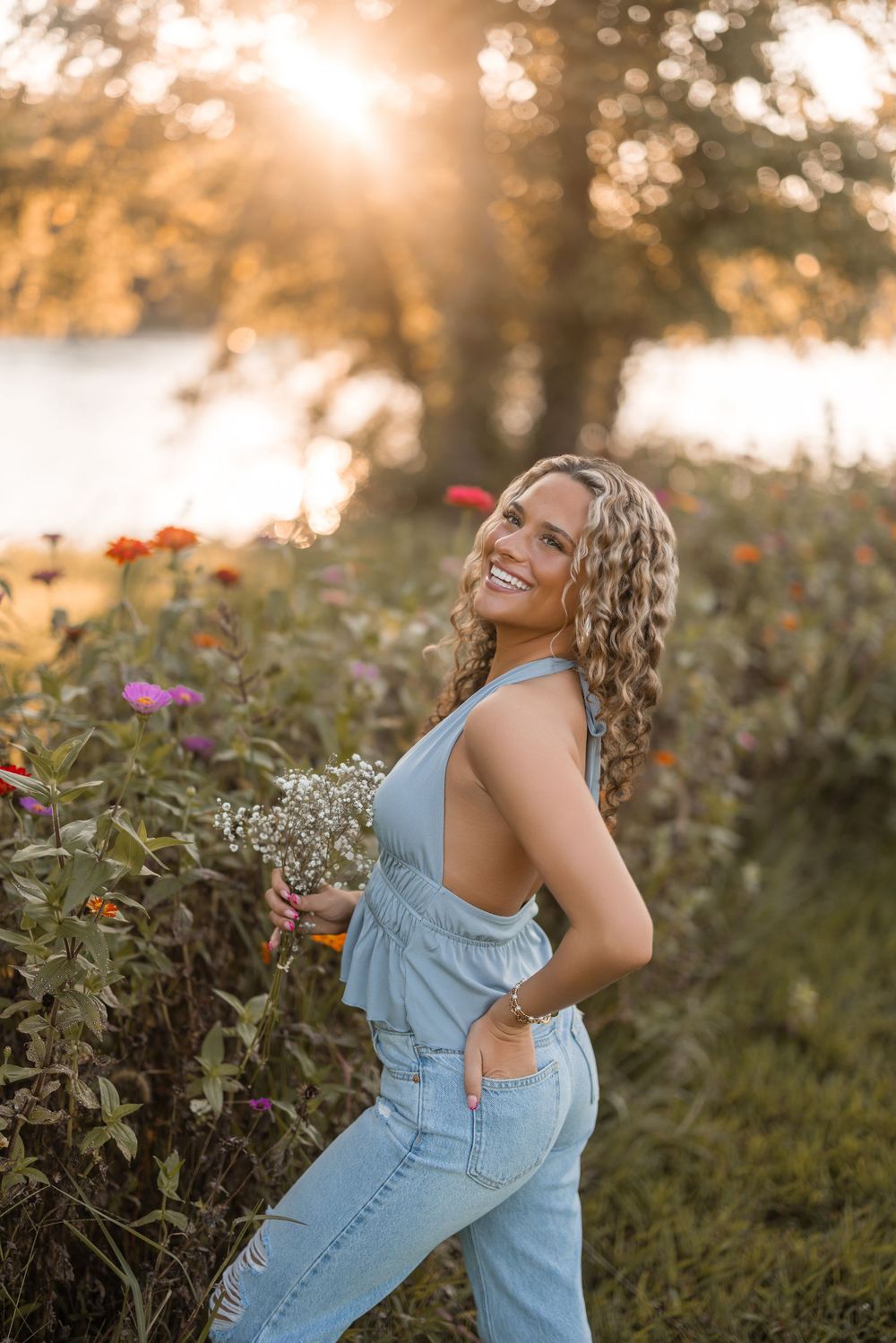 Person in light denim overalls poses playfully in a wildflower garden during golden hour sunset.