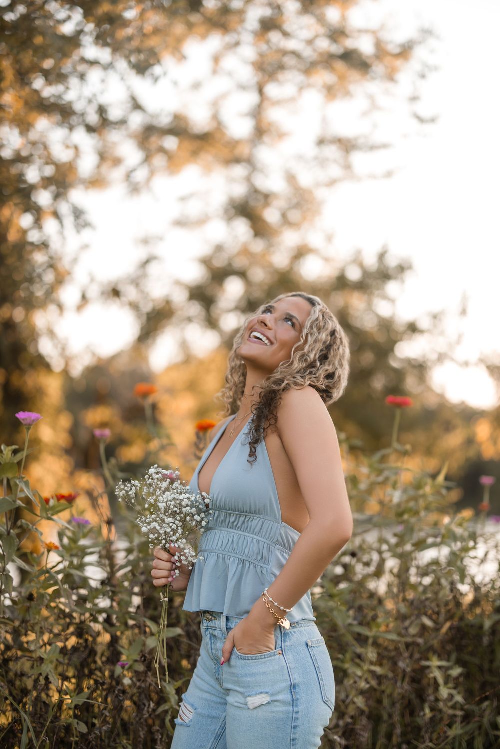 A woman laughs joyfully in a wildflower field at sunset wearing a light blue top and holding delicate flowers.
