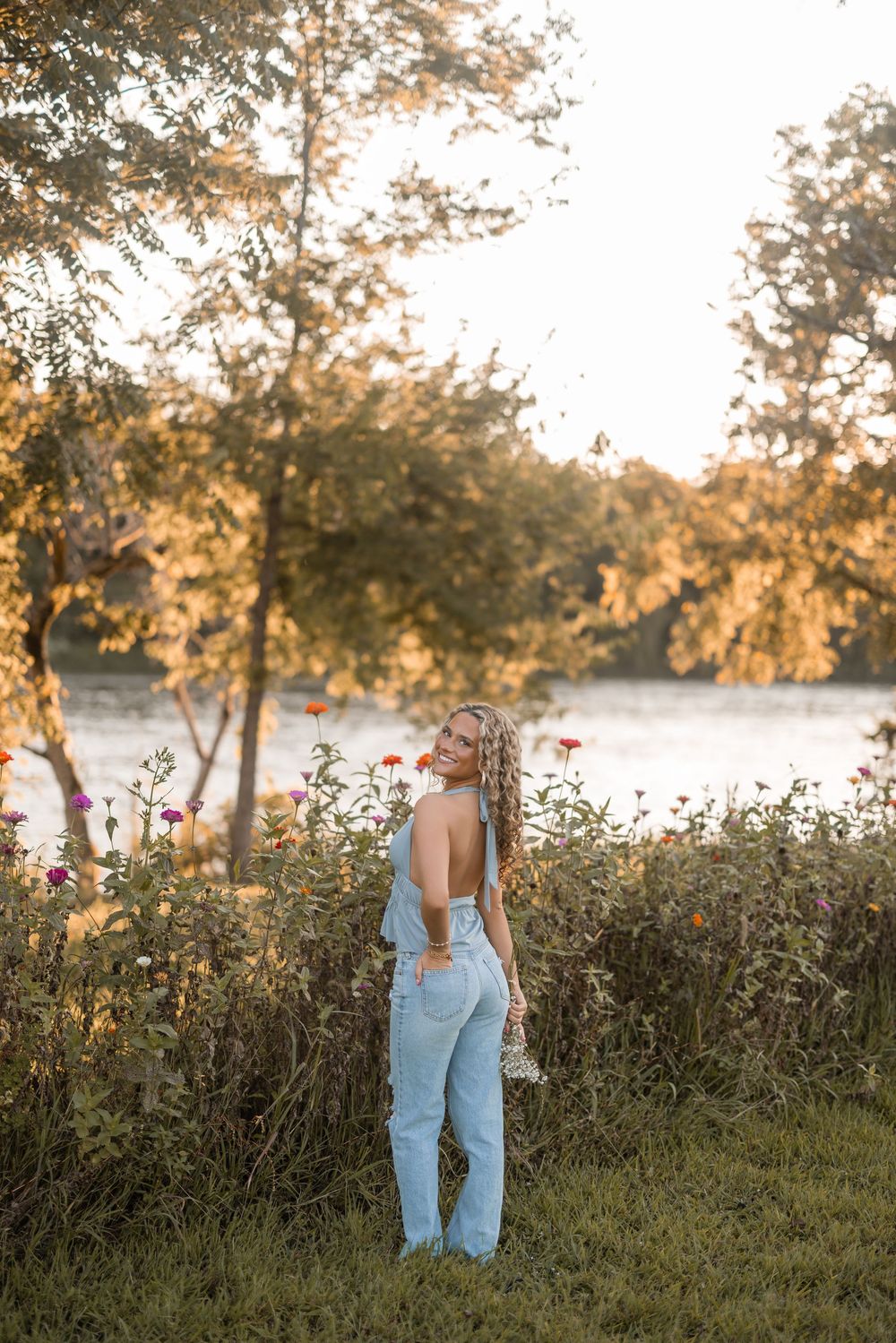 Figure in denim overalls stands among wildflowers against a sunlit lake backdrop.