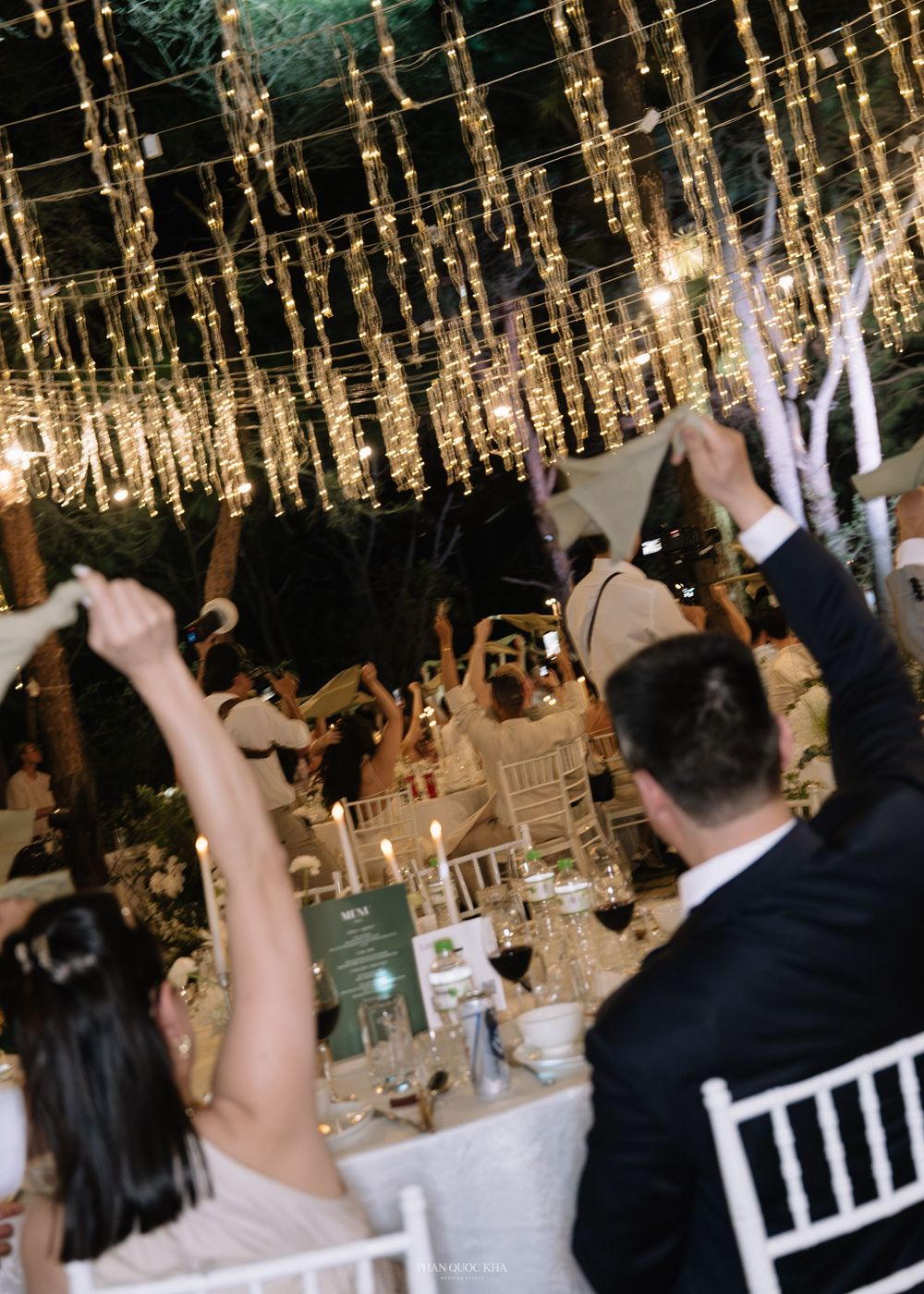 Excited wedding guests wave napkins and phones in the air during a lively outdoor evening reception.