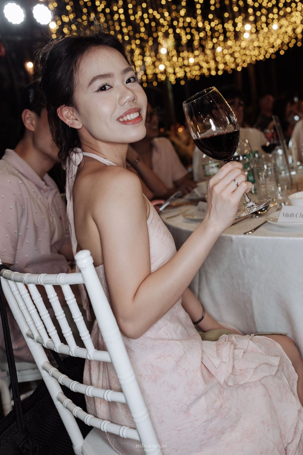 Smiling young woman in a pink dress raises a glass of red wine at an outdoor wedding reception.
