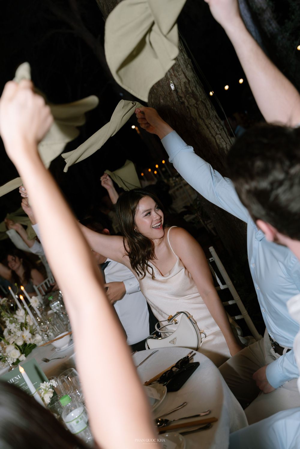 Laughing wedding guest waves a napkin in the air during a lively outdoor evening reception celebration.