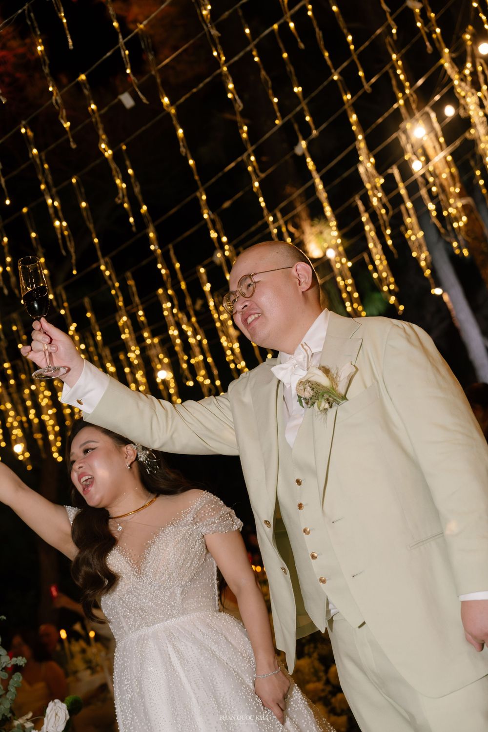 Joyful bride and groom in cream suit celebrating under golden fairy lights at elegant wedding reception.