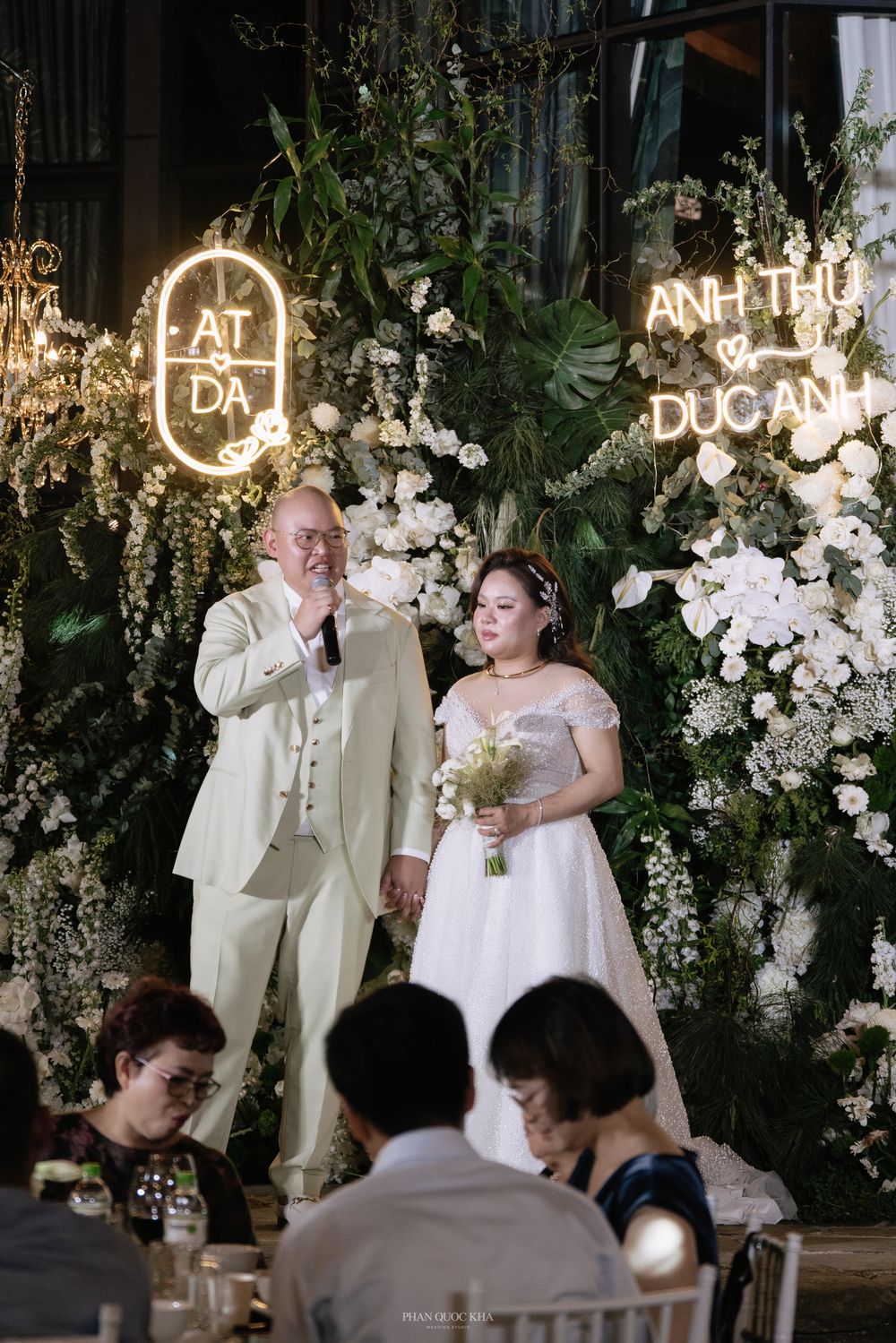 Groom speaking at wedding reception with bride beside floral arch and neon monogram sign.