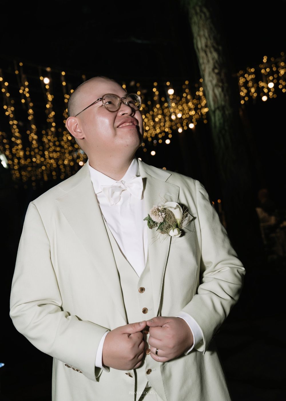 Groom in ivory suit with boutonniere smiling under string lights at outdoor wedding reception.