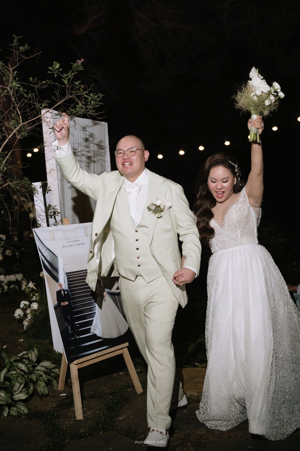 Bride and groom celebrating joyfully at outdoor wedding with piano backdrop and string lights.