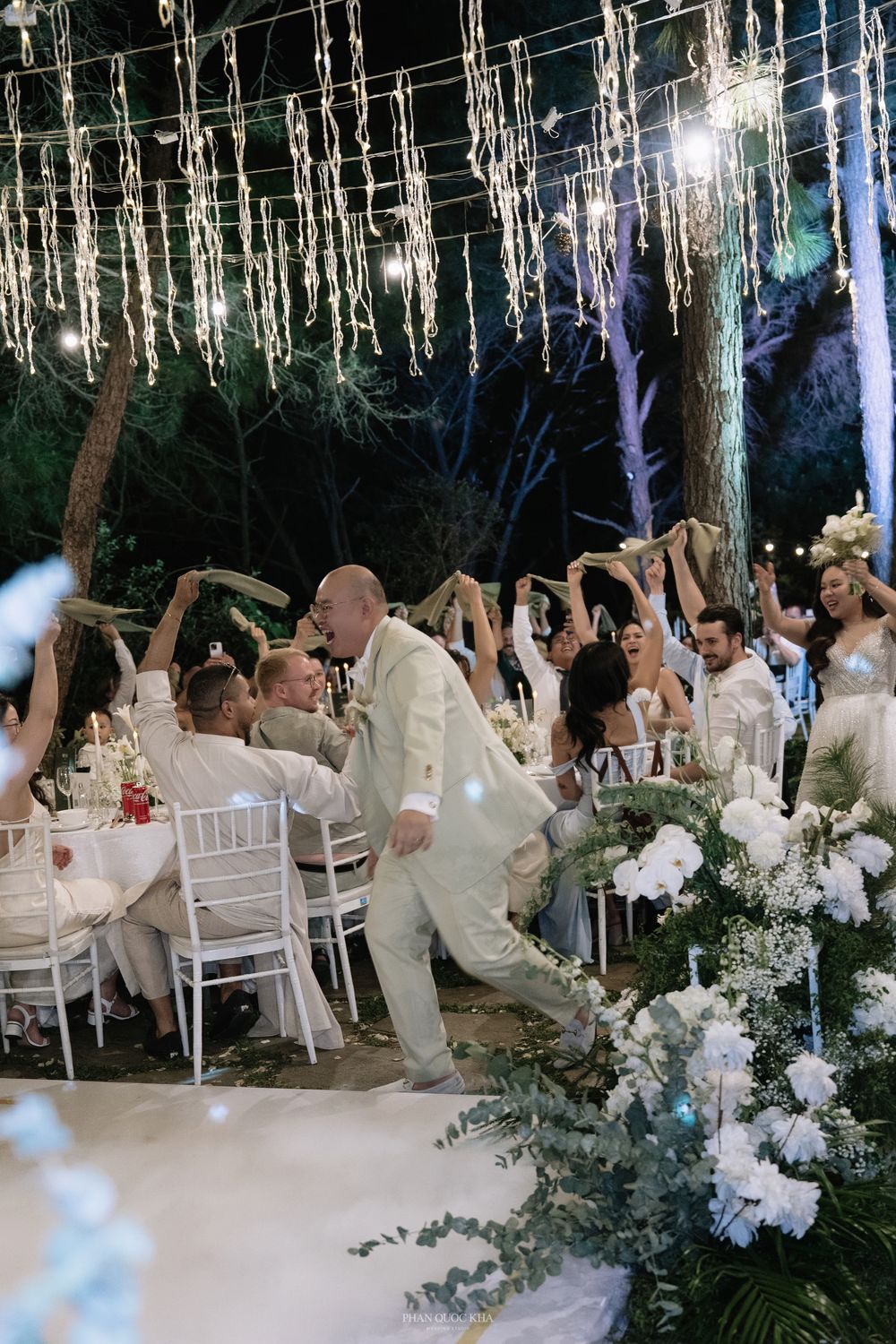 Wedding guests raising arms in celebration at elegant outdoor reception with string lights.