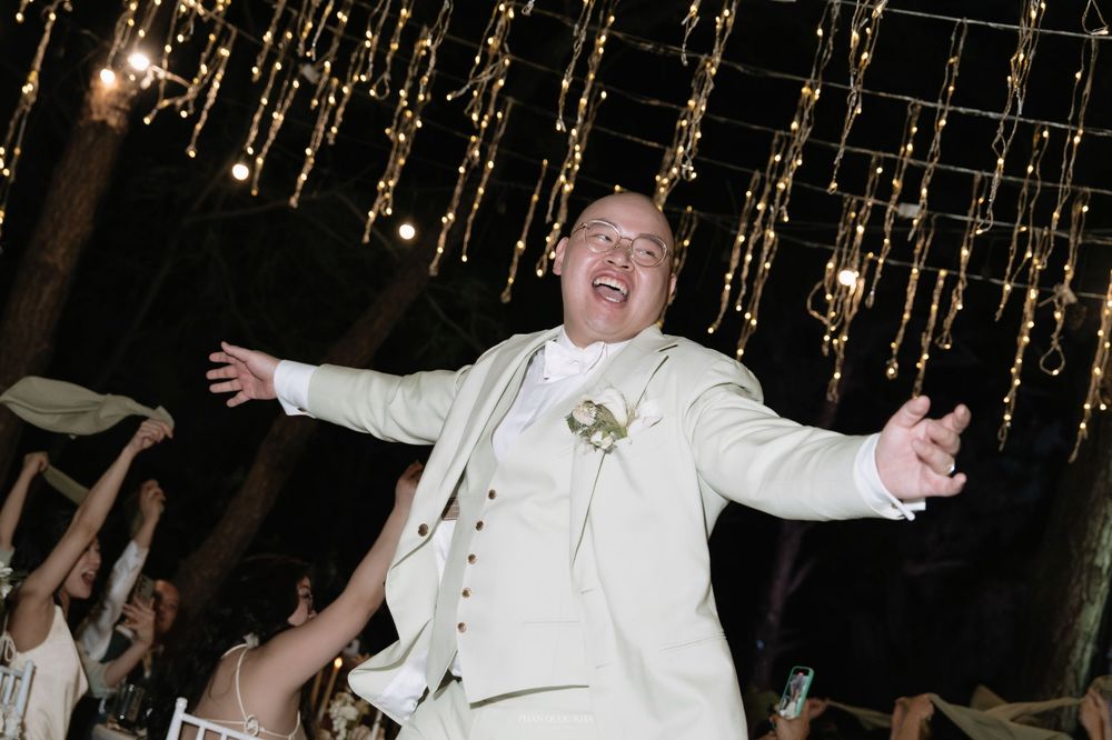 Groom in ivory suit celebrating with arms wide open on wedding dance floor with guests.