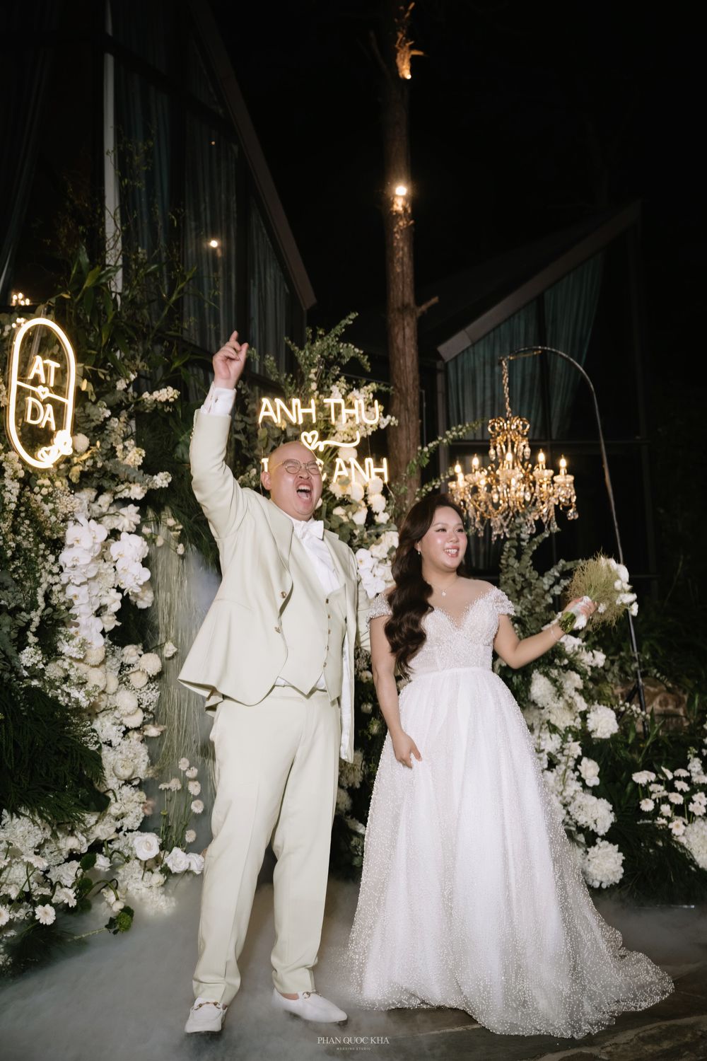 Bride and groom celebrating in front of floral arch with neon wedding sign at night.