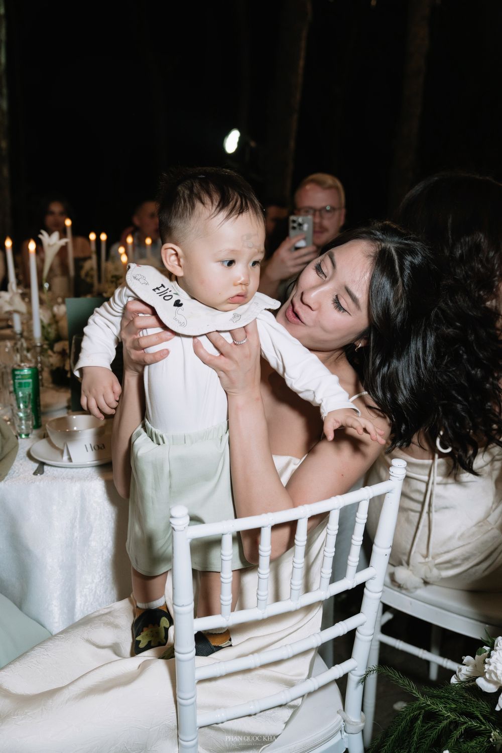 Mother in white dress holding baby at elegant candlelit wedding reception table.