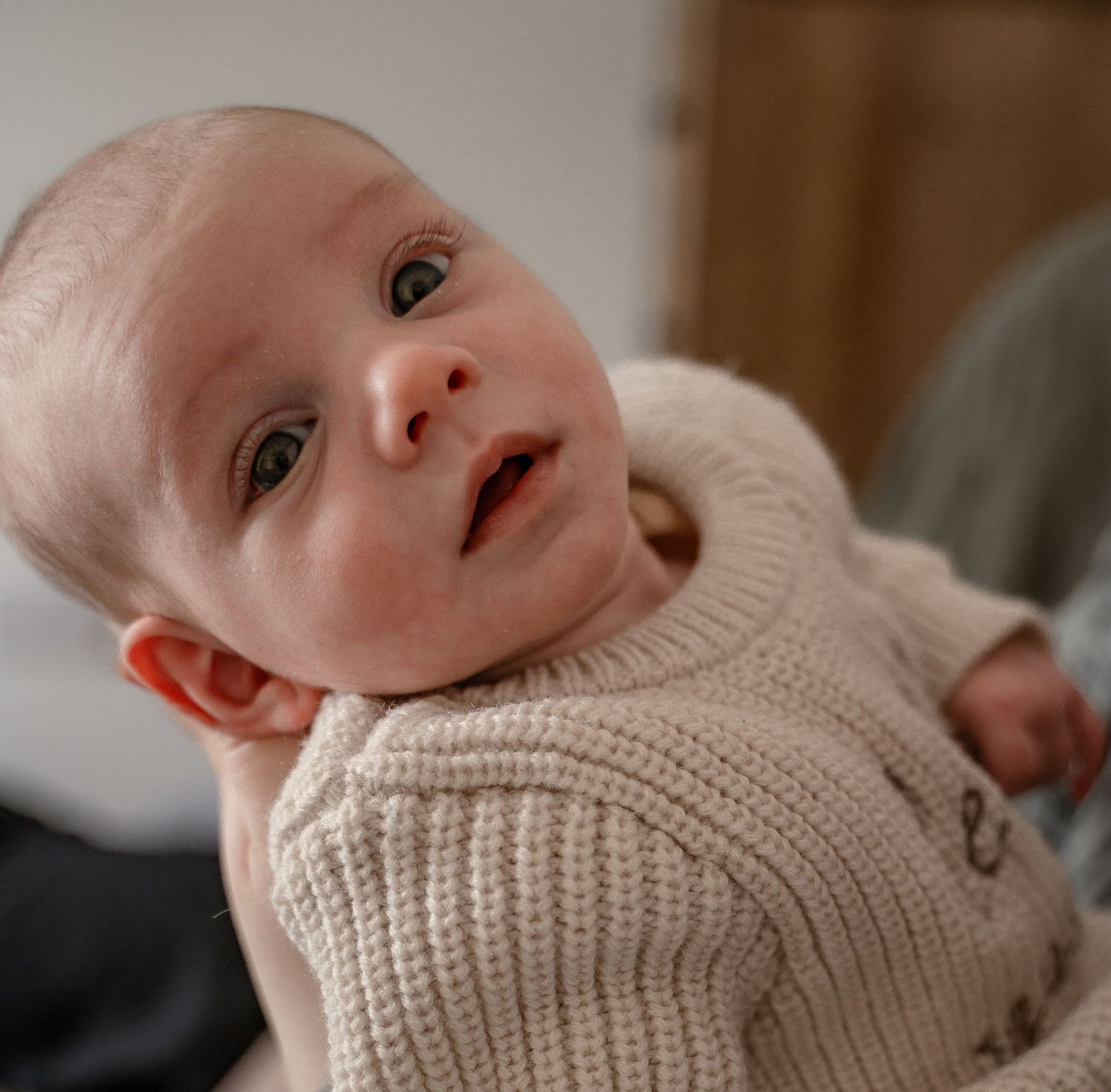 Baby wearing a cozy beige knit sweater gazes upward with bright eyes and a gentle expression.
