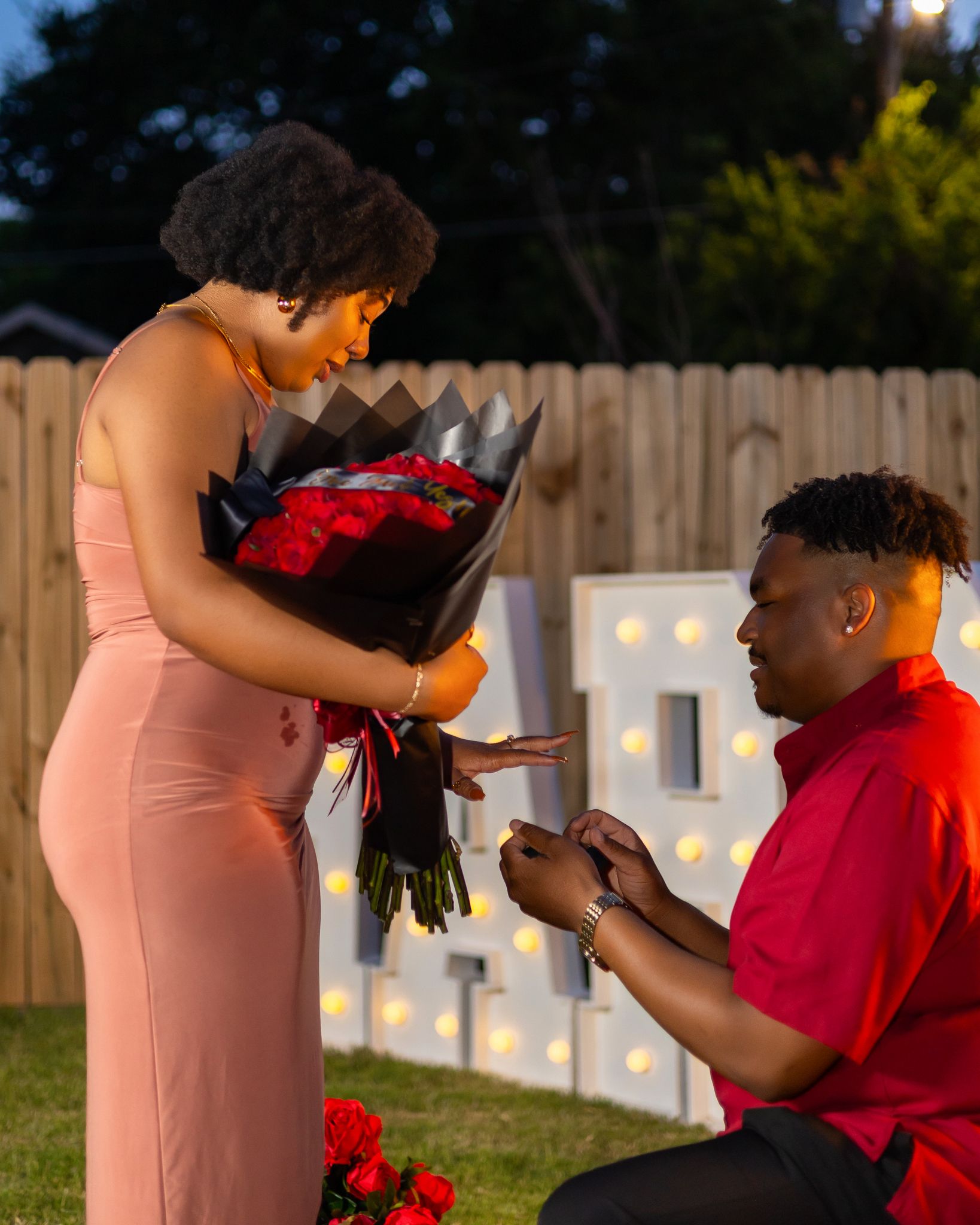Evening engagement photoshoot with romantic lighting and red roses in a backyard setting.