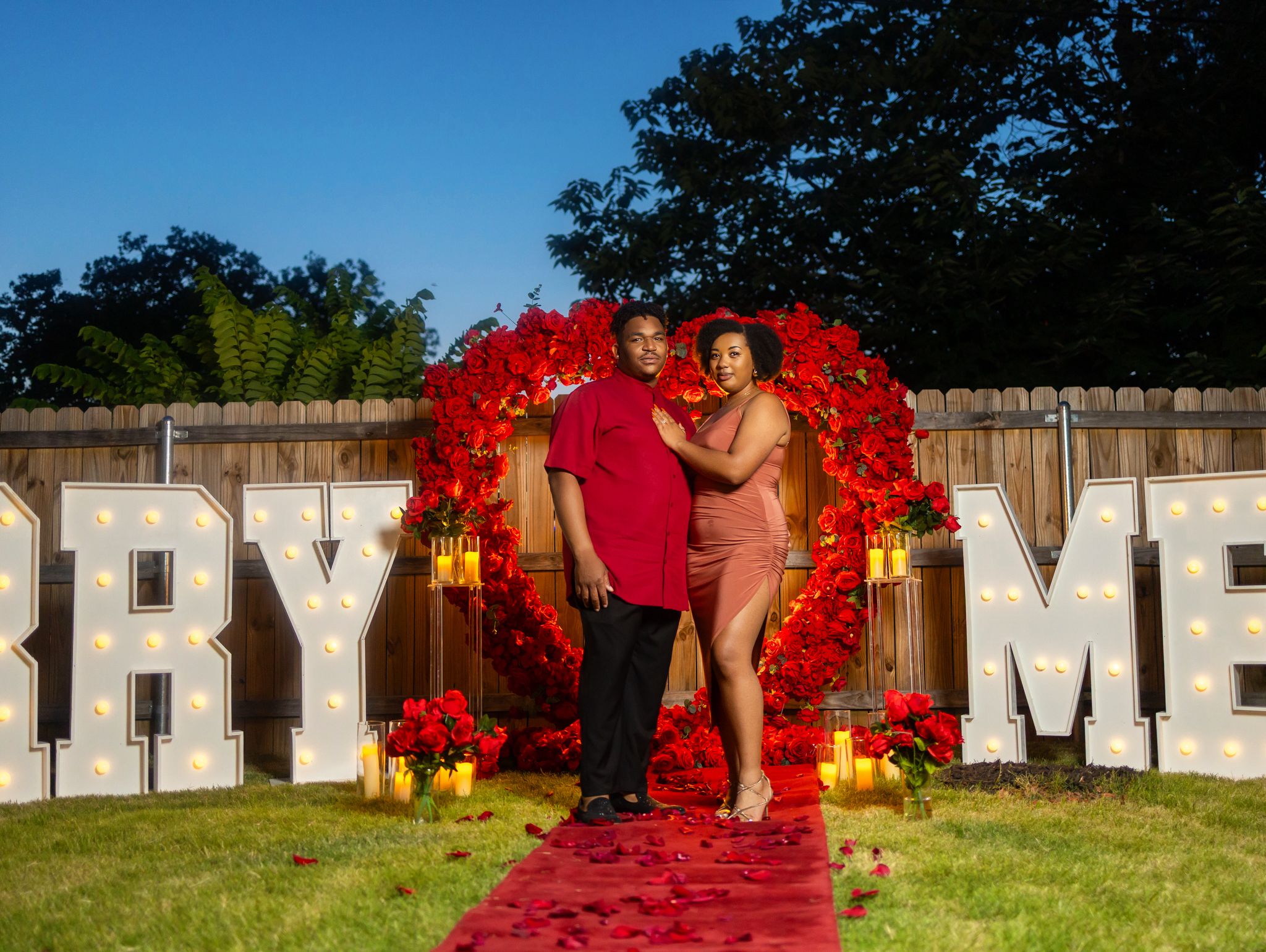 Large white letters spelling out 'MARRY ME' decorated with red flowers in an outdoor setting.
