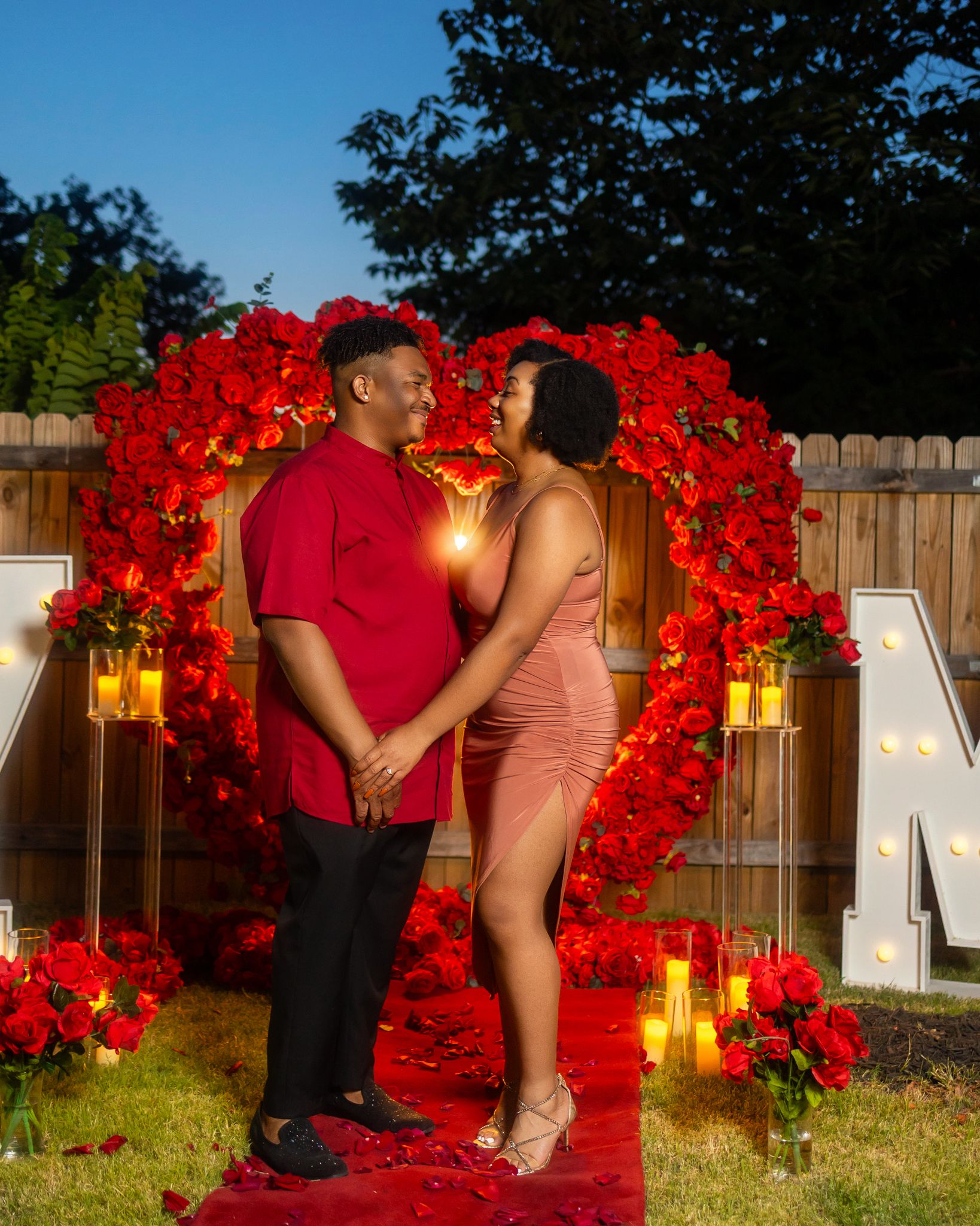 A couple poses together near decorative red flowers and illuminated letters during a proposal.