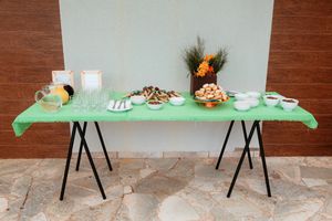 Long table with green tablecloth displays coffee and snacks with black chairs arranged along the sides against a brick wall.