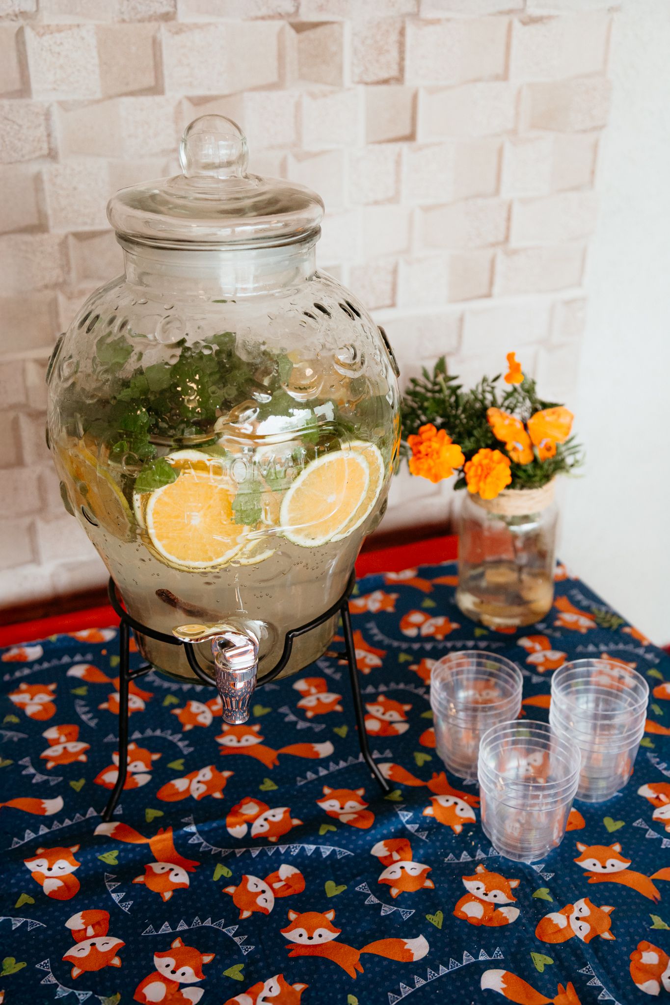 A glass beverage dispenser filled with citrus infused liquid stands on a navy fox-patterned tablecloth.