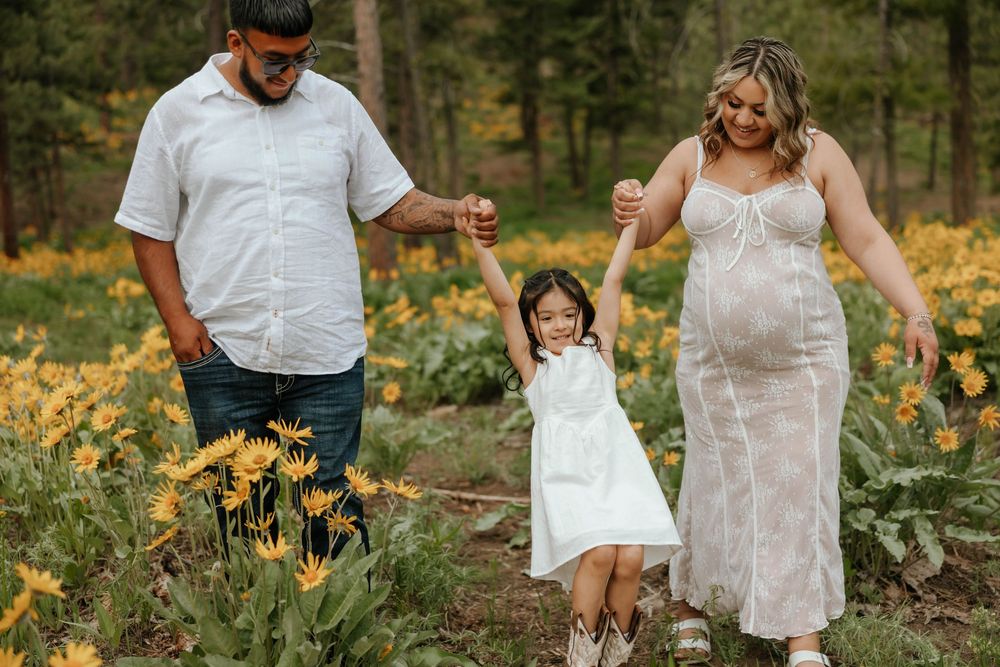 Family maternity photoshoot among yellow wildflowers and pine trees.