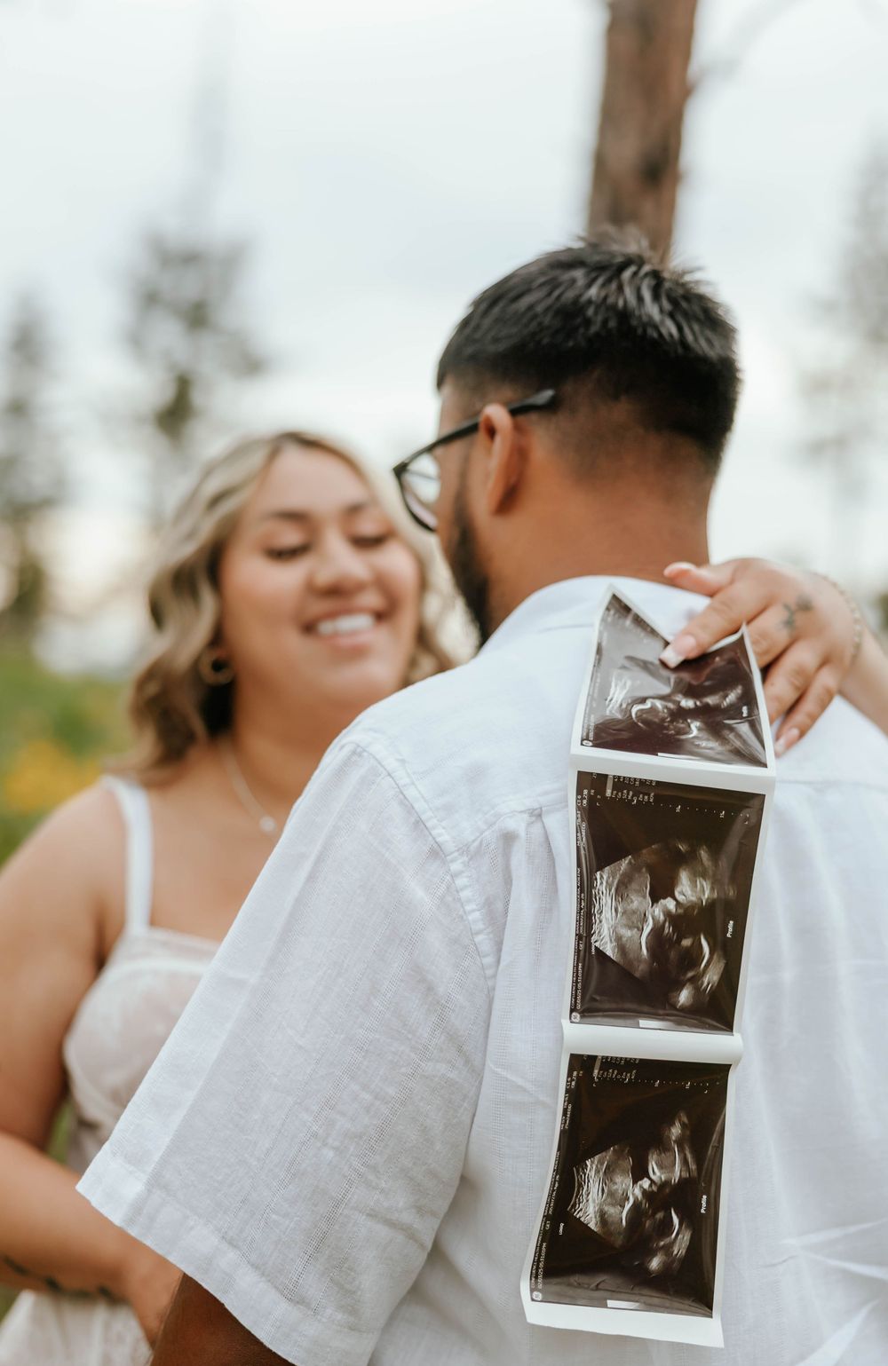 Couple sharing tender moments while holding ultrasound photos outdoors.