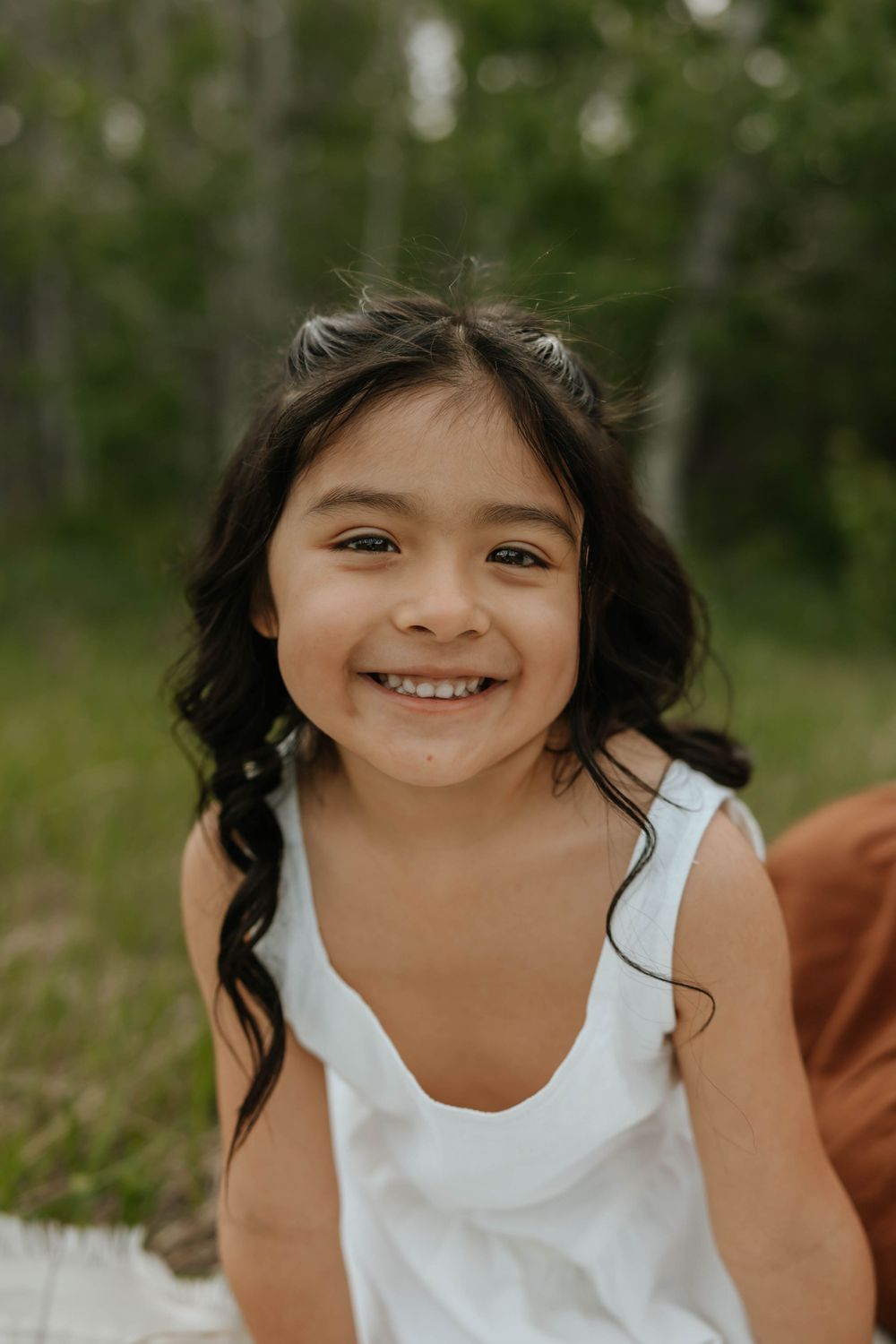 A smiling child in a white dress plays outdoors in a natural setting with trees in the background.
