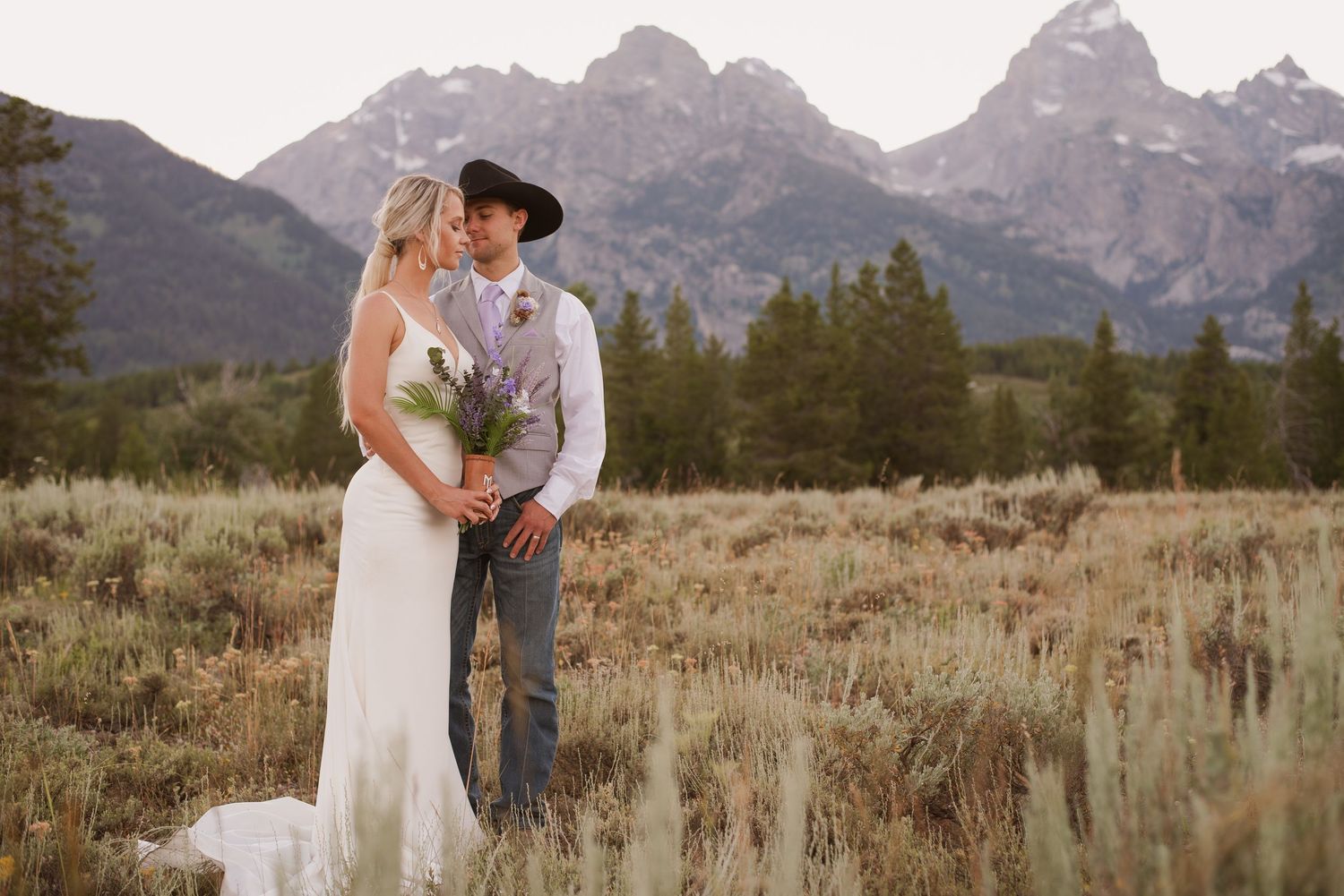 Wedding couple standing in field with dramatic mountain landscape backdrop.