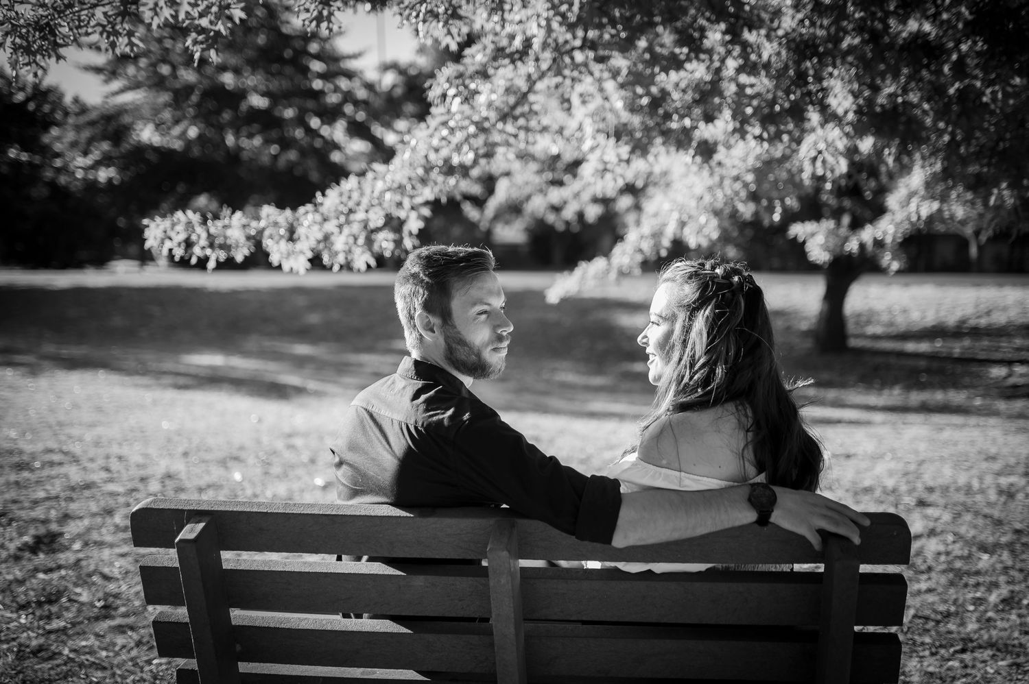 A couple sits together on a park bench in black and white, sharing an intimate moment in nature.