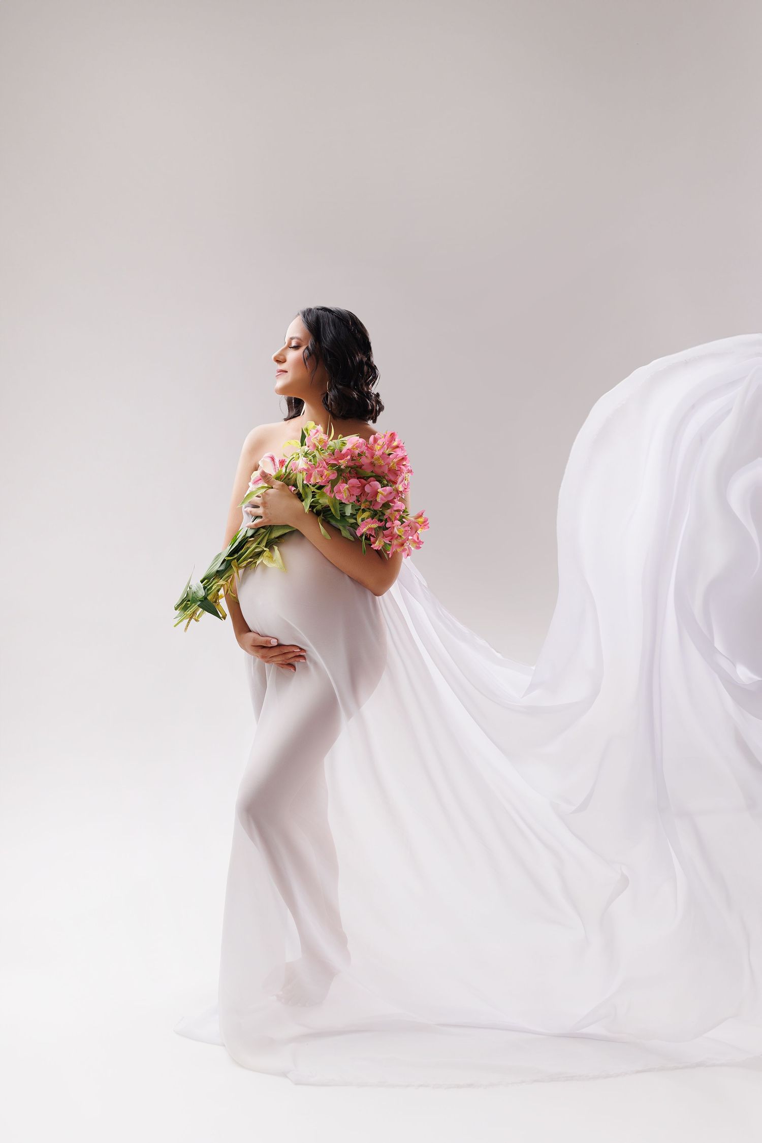 Elegant maternity portrait with flowing white fabric and pink flowers against light backdrop.