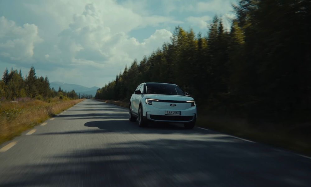 Rural mountain road curves through forested landscape with white pickup truck in distance.