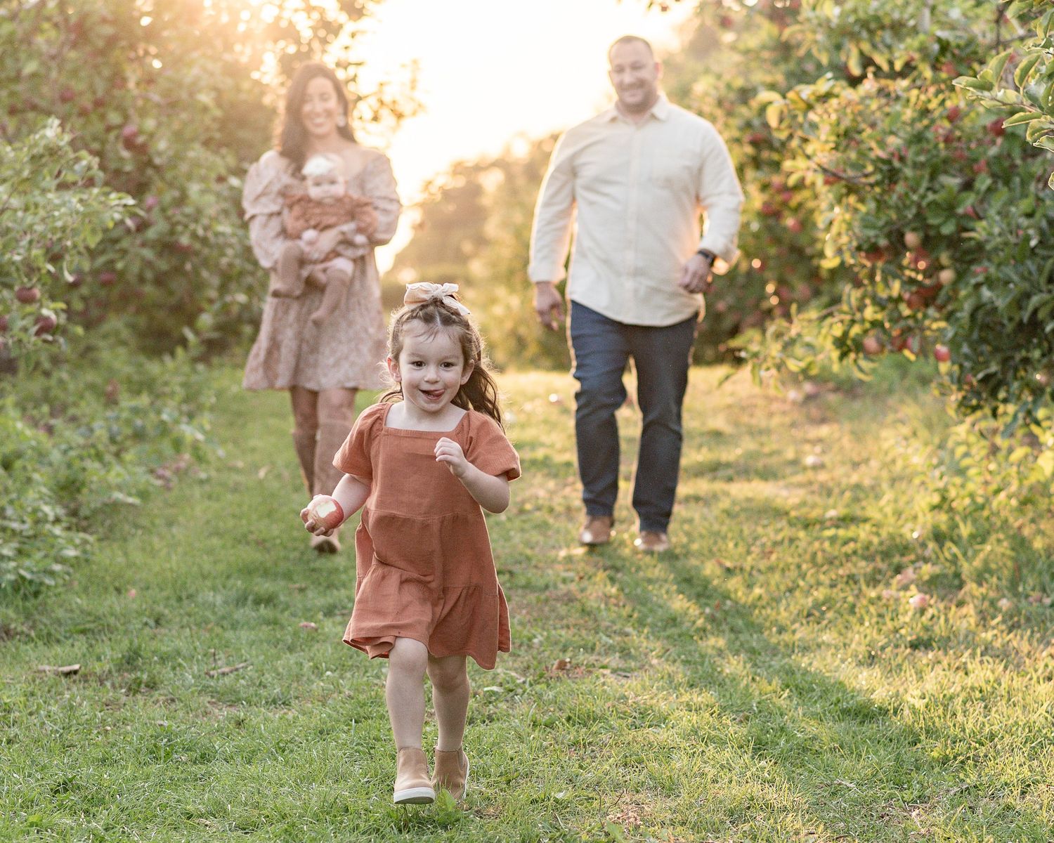 A young family of four showing a small child in a rust-colored dress walking through a sunlit meadow at golden hour.