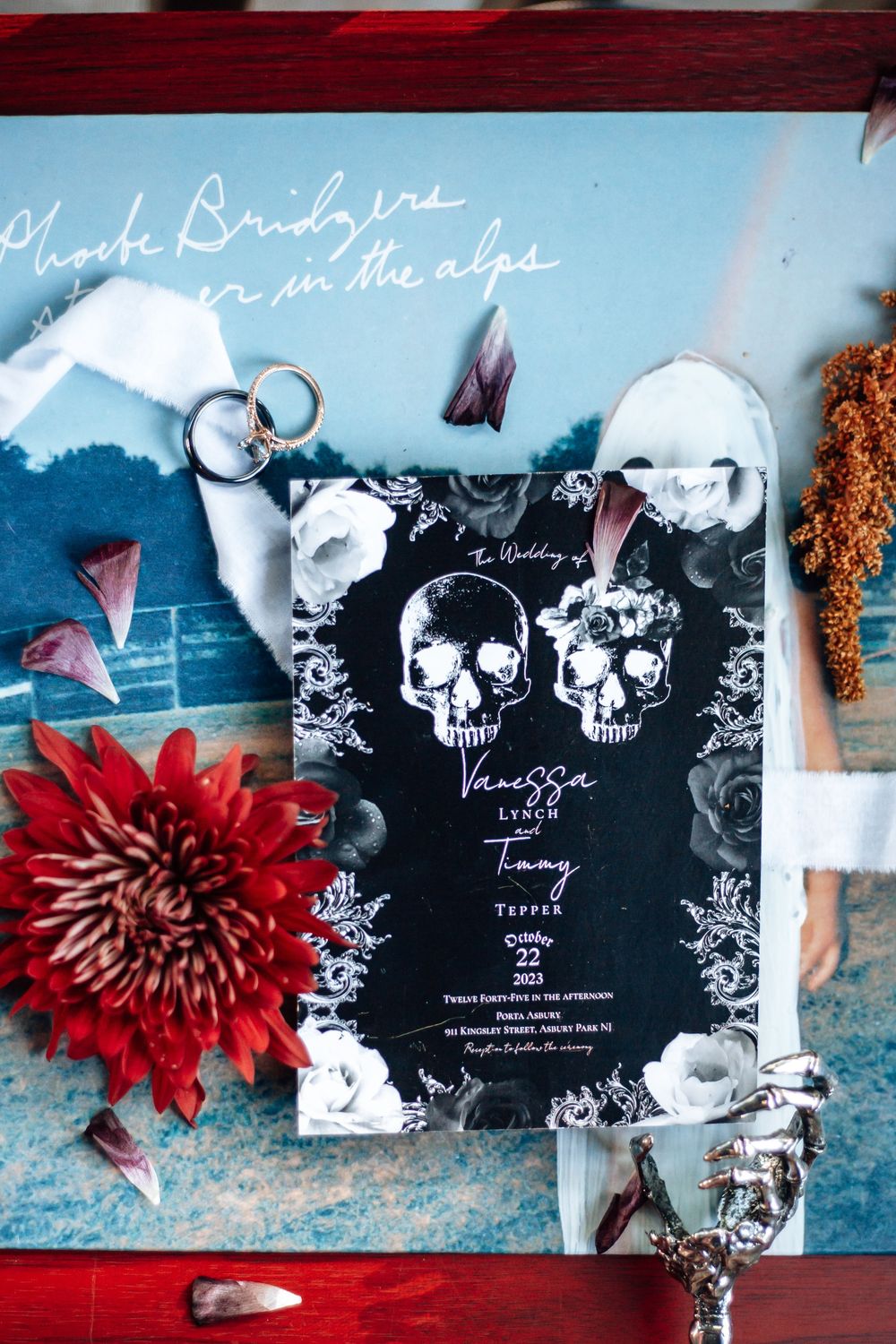 Gothic wedding invitation cards in black with skull designs arranged with red dahlias against a blue background.