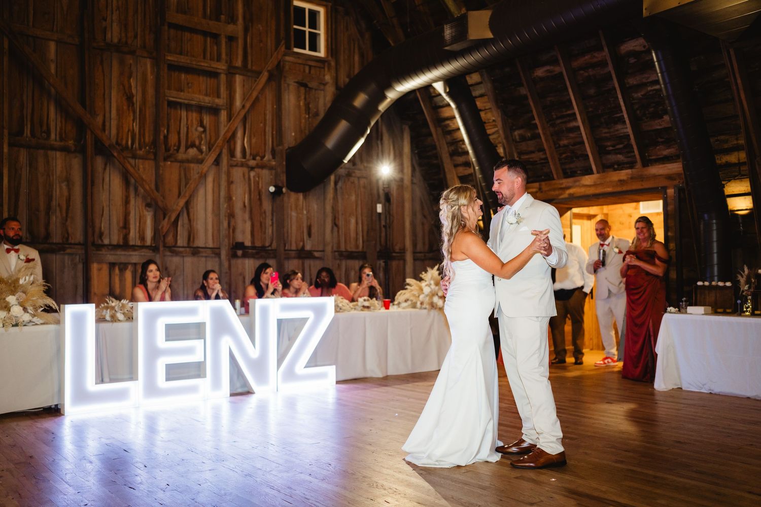 A rustic barn wedding venue with illuminated chandelier and couple sharing their first dance on wooden floor beneath.