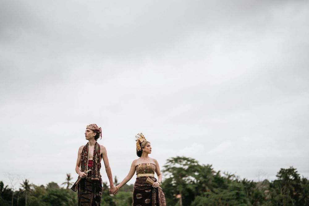 Couple in traditional Balinese attire holding hands against cloudy sky and tropical landscape.