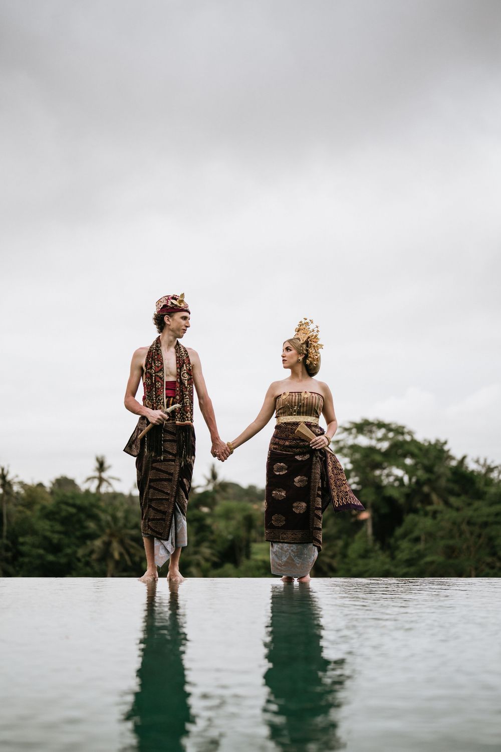 Couple in traditional Indonesian dress stands at infinity pool edge with tropical forest view.