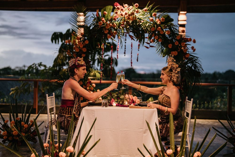 Couple dining at romantic table setting under floral arch with tropical backdrop.