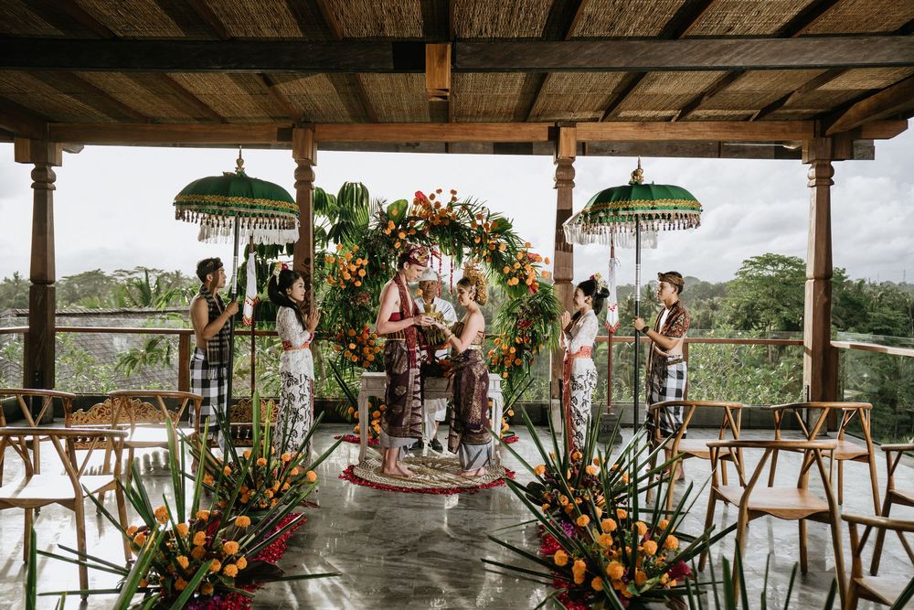 Tropical wedding ceremony setup with floral arch and umbrellas on wooden deck overlooking jungle.