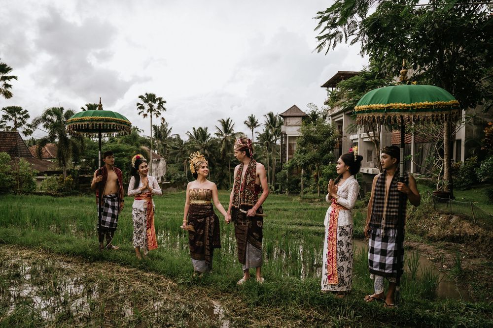 Wedding party in traditional Balinese attire poses in tropical garden setting with palm trees.