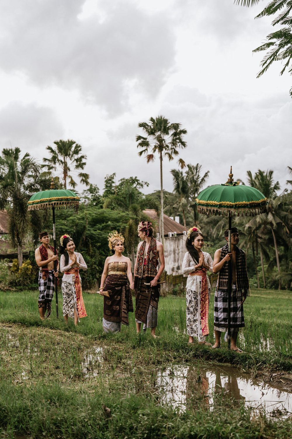 A wedding party poses in a lush tropical garden setting with palm trees and green umbrellas.
