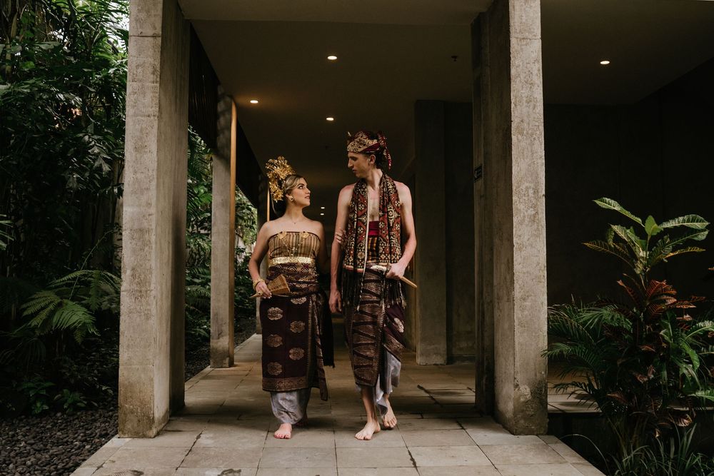 Couple in traditional Indonesian clothing poses in modern colonnade with tropical garden backdrop.