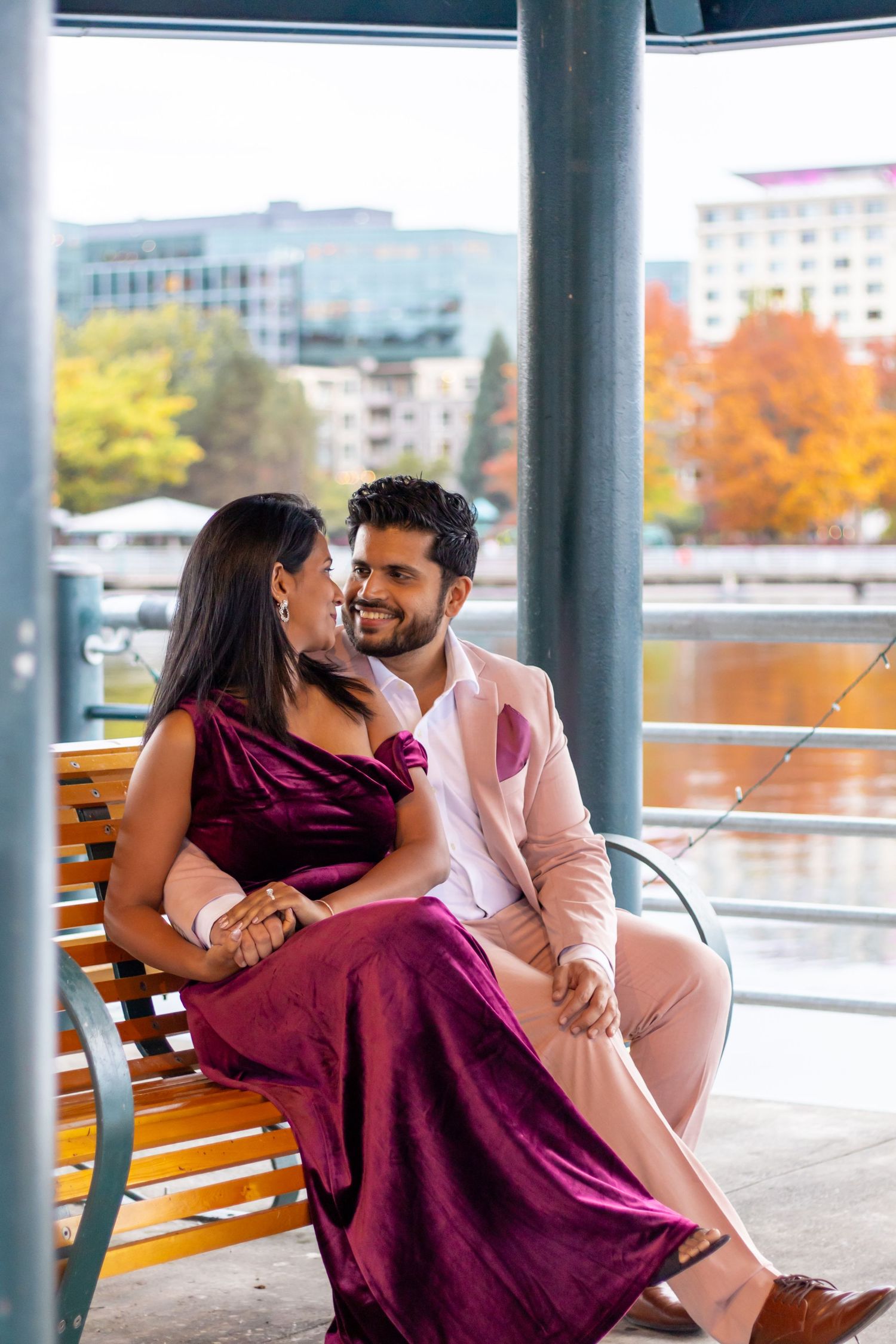 An elegant couple in formal attire sits on a bench sharing a romantic moment with a city waterfront backdrop in autumn.