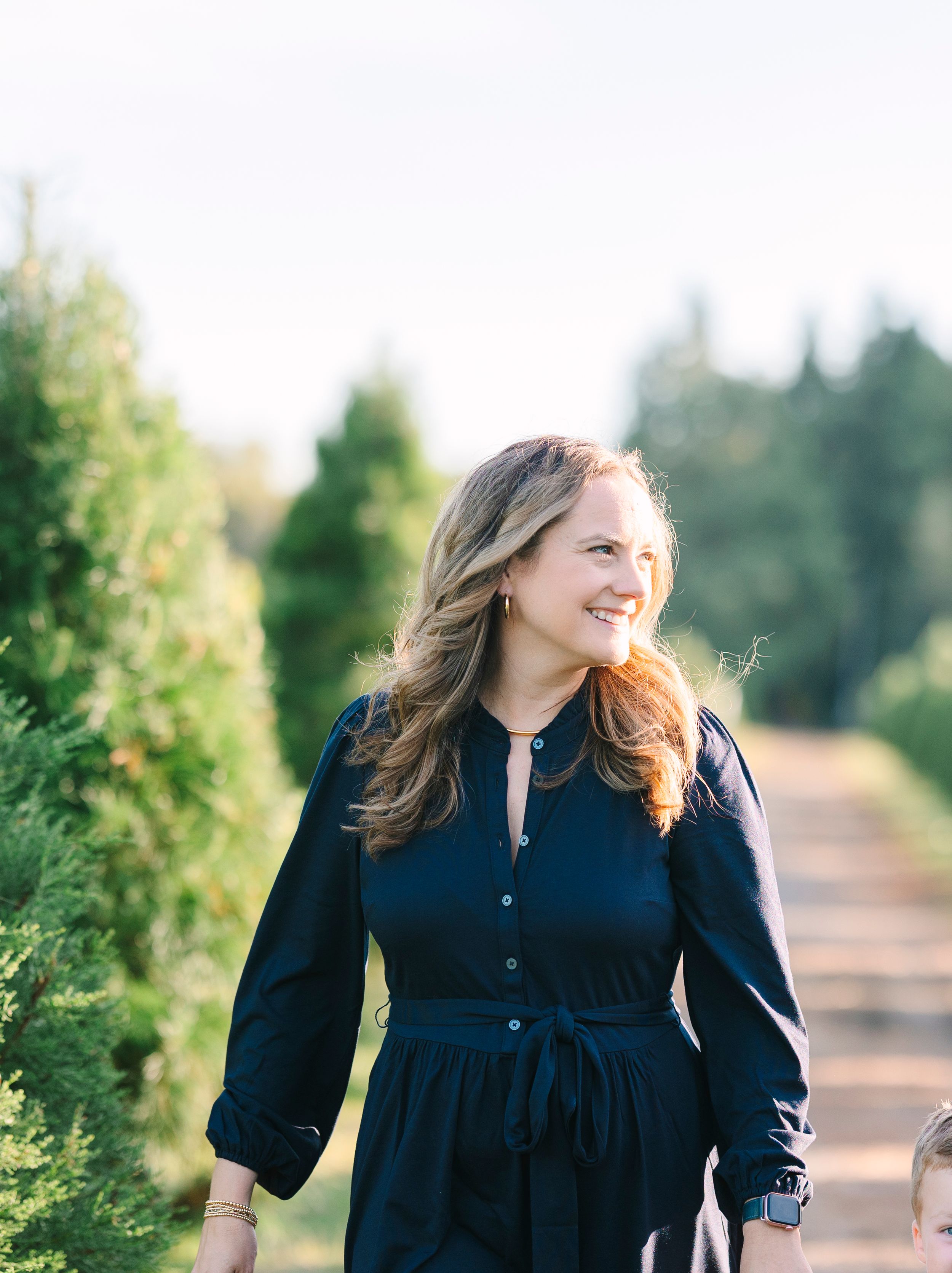 A person in a navy blue dress stands in a sunlit path between evergreen trees.