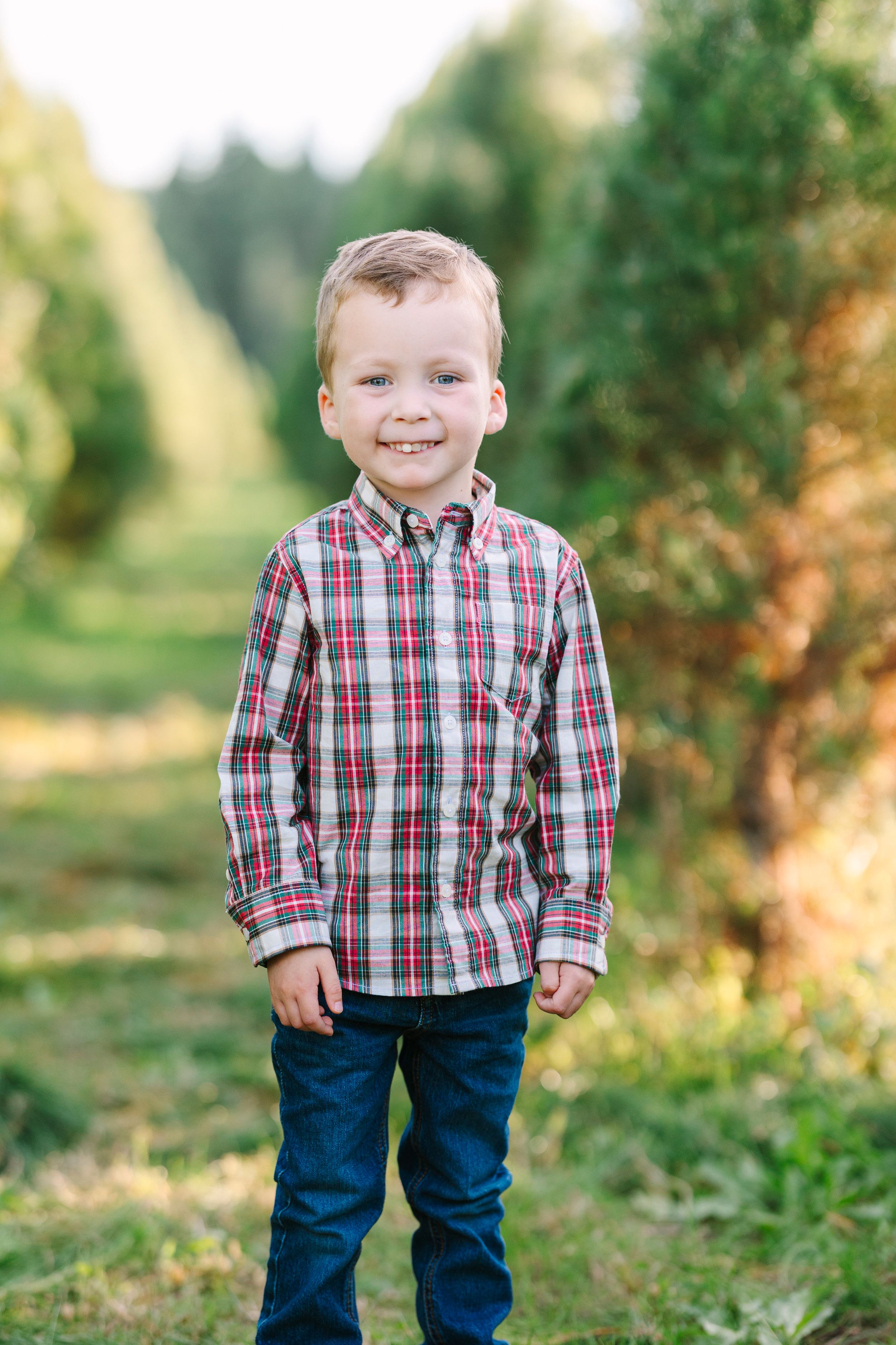 A child in a plaid shirt and jeans stands smiling in a green outdoor setting.