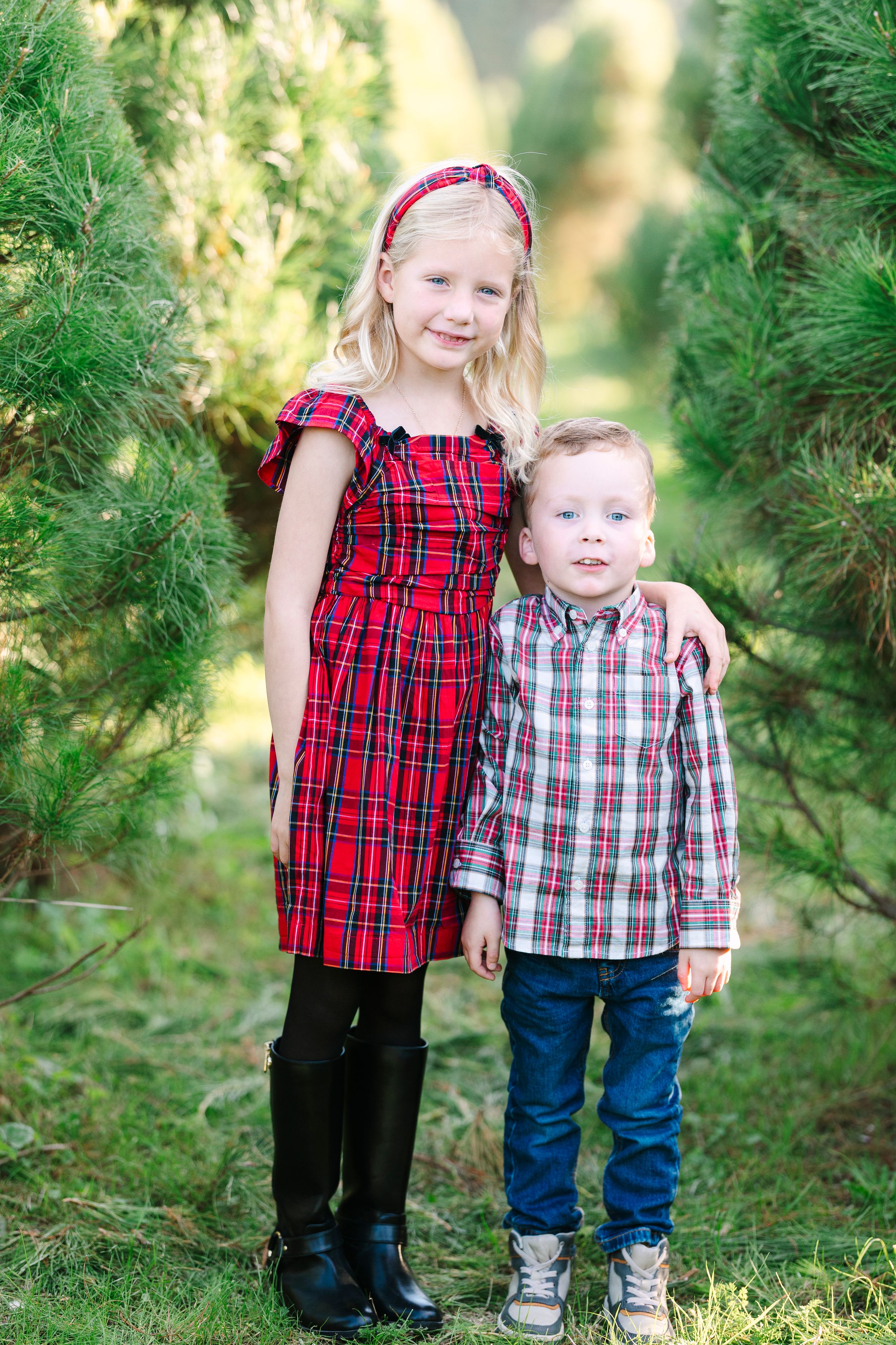 Two children in coordinating plaid outfits stand together among evergreen trees.