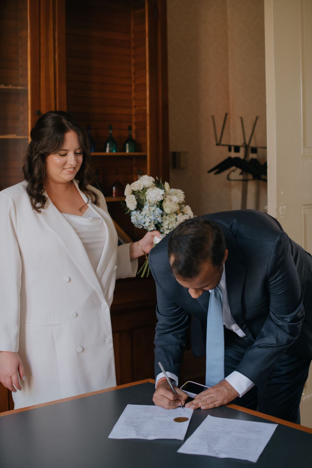 Two people in formal attire sign documents at a wooden desk with floral arrangements in the background.